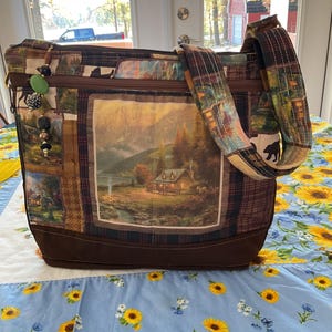 May include: A brown quilted tote bag with a zipper closure and a long adjustable strap. The bag features a patchwork design with various nature-themed prints, including a cabin in the woods, a bear, and trees. The bag has a brown canvas bottom and a gold zipper pull.