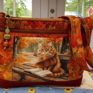 Op de afbeelding: Gewatteerde draagtas met een herfstbladerenpatroon in tinten oranje, rood en geel. Het middenpaneel toont een schilderij van een rode kat met een sjaal die een boek leest op een parkbank. De tas heeft een rode basis en bijpassende handvatten.