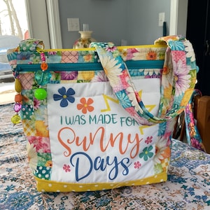 May include: A colorful tote bag with a floral patchwork design. The bag features a white panel with the text "I WAS MADE FOR Sunny Days" in orange and blue. It has a yellow trim and a teal zipper.