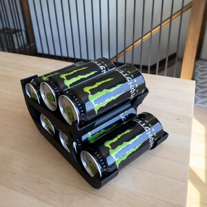 May include: A black, tiered can organizer holding six Monster Energy drinks. The cans are black with a green and yellow claw design. The organizer sits on a light wood surface.