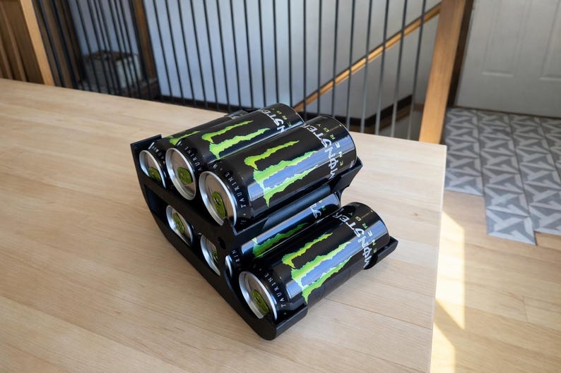 May include: A black, tiered can organizer holding six Monster Energy drinks. The cans are black with a green and yellow claw design. The organizer sits on a light wood surface.