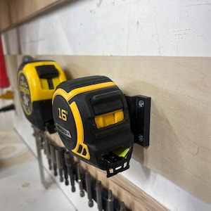 May include: Two yellow and black tape measures are mounted on a wooden wall. The tape measure on the right has a black handle and a yellow body with the text "16'" printed on it.