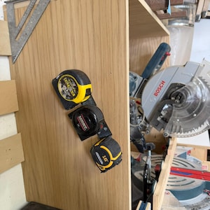 May include: A woodworking shop scene featuring a wooden cabinet with three tape measures attached. A silver and blue circular saw is visible, along with a metal speed square. The cabinet is made of light-colored wood.
