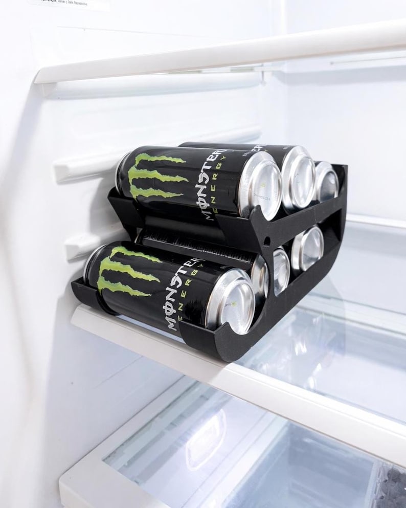 May include: A black can organizer holding energy drink cans inside a refrigerator. The cans are black with a green claw design and the word "Monster" in white. The organizer is on a shelf.