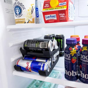 May include: A refrigerator interior with various food items. Includes a container of Greek yogurt, Lactaid milk, energy drinks, protein drinks, and a carton of eggs. A black can organizer holds the energy drinks.