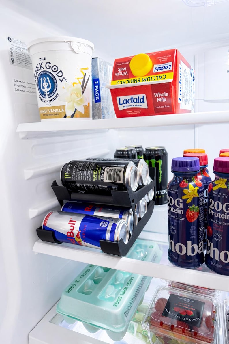 May include: A refrigerator interior with various food items. Includes a container of Greek yogurt, Lactaid milk, energy drinks, protein drinks, and a carton of eggs. A black can organizer holds the energy drinks.