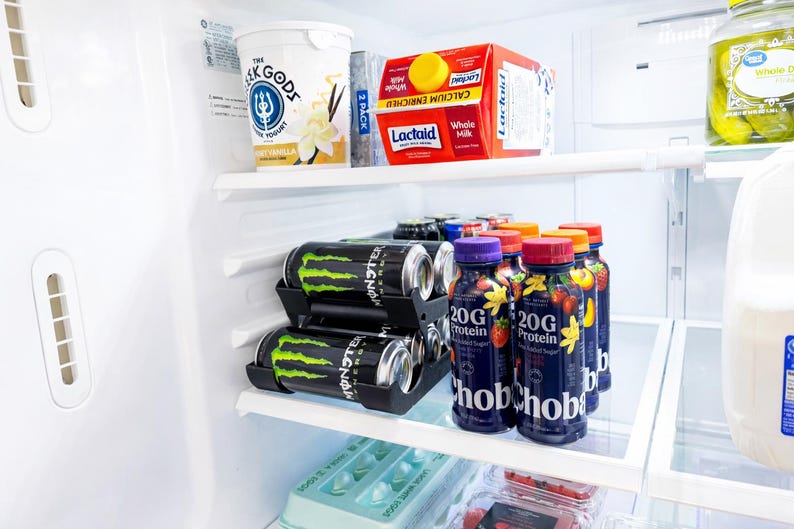 May include: A refrigerator interior with various food items. Includes yogurt, Lactaid milk, energy drinks, and protein shakes. The energy drinks are black with green accents, and the protein shakes are in dark blue bottles with fruit graphics.