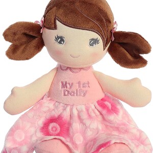 May include: A pink and white plush doll with brown hair in pigtails. The doll is wearing a pink dress with a floral pattern and has the words "My 1st Dolly" embroidered on the front.