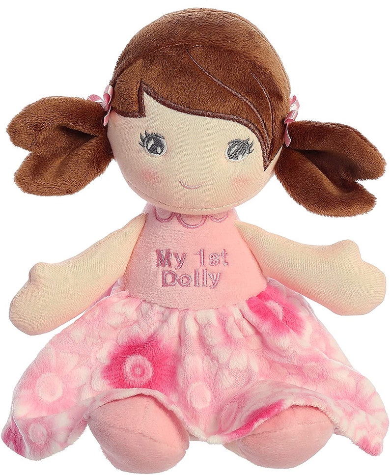 May include: A pink and white plush doll with brown hair in pigtails. The doll is wearing a pink dress with a floral pattern and has the words "My 1st Dolly" embroidered on the front.