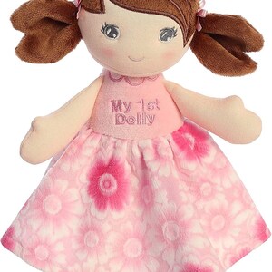 May include: A pink and white floral dress with a brown haired doll wearing pink shoes. The doll has embroidered eyes and a smile. The dress has the text "My 1st Dolly" embroidered on the front.