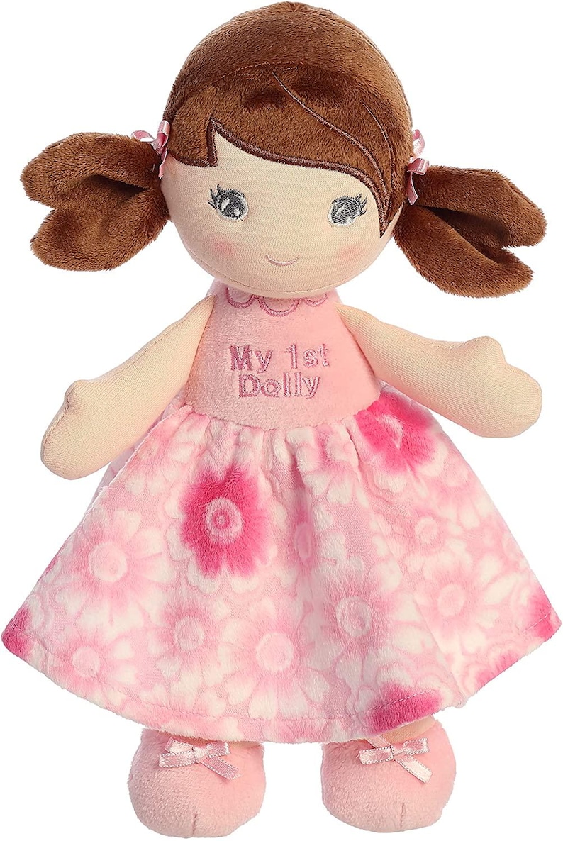 May include: A pink and white floral dress with a brown haired doll wearing pink shoes. The doll has embroidered eyes and a smile. The dress has the text "My 1st Dolly" embroidered on the front.