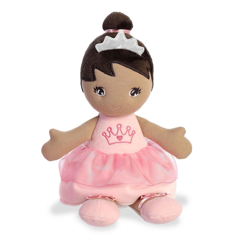 May include: A brown-skinned plush doll wearing a pink ballerina dress with a crown and a heart on the front. The doll has brown hair and a silver tiara.