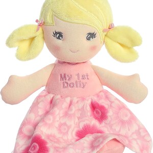 May include: A pink and white plush doll with yellow hair in pigtails. The doll is wearing a pink dress with a floral pattern and has the words "My 1st Dolly" embroidered on the front.