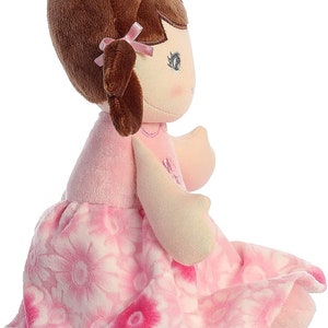 May include: A pink and white floral dress doll with brown hair and a pink bow. The doll is sitting with its legs crossed.