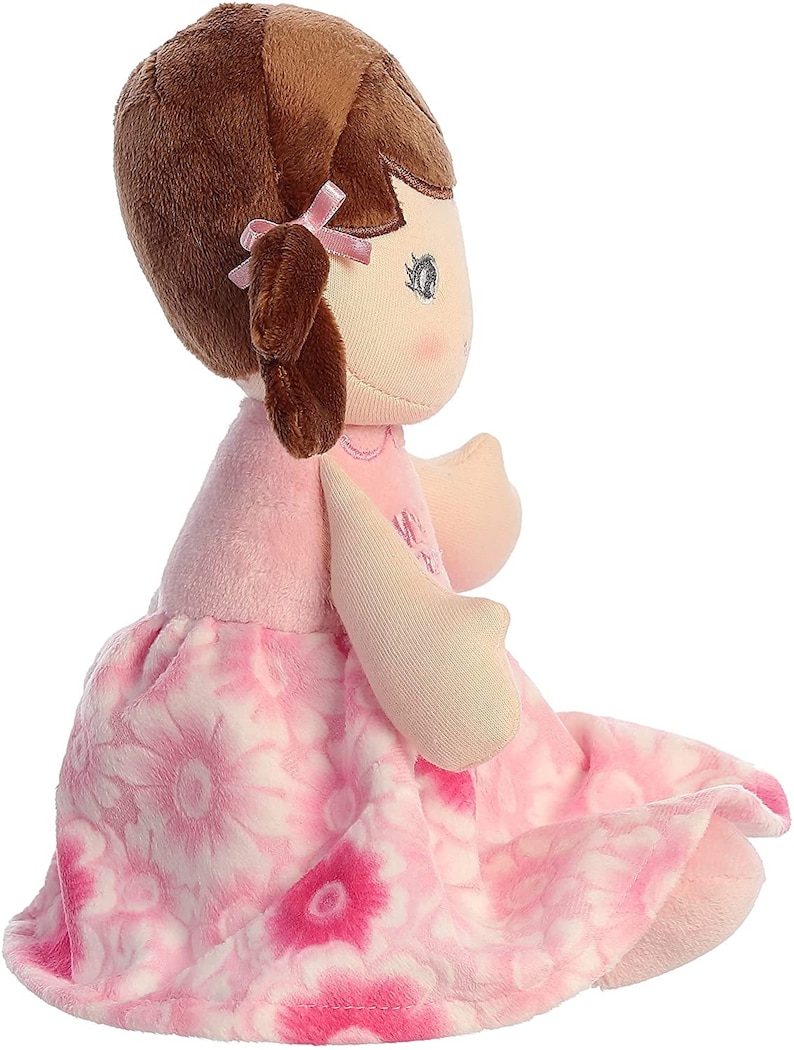 May include: A pink and white floral dress doll with brown hair and a pink bow. The doll is sitting with its legs crossed.