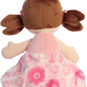 May include: A pink plush doll with brown hair in pigtails wearing a pink dress with white flowers.