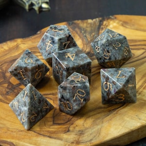 May include: A set of seven speckled gray and gold polyhedral dice for tabletop games, displayed on a wooden board. A miniature figurine and two metal coins are also present. A sword is visible in the background.
