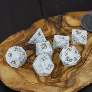 May include: A set of seven white marble dice with gold numbers on a wooden tray. The dice are a d4, d6, d8, d10, d12, and two d20s.