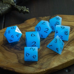 Puede incluir: Un conjunto de siete dados azules que brillan en la oscuridad con números negros. Los dados están sobre una superficie de madera.