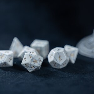 May include: A set of white marble dice with gold numbers. The dice are on a dark blue surface.