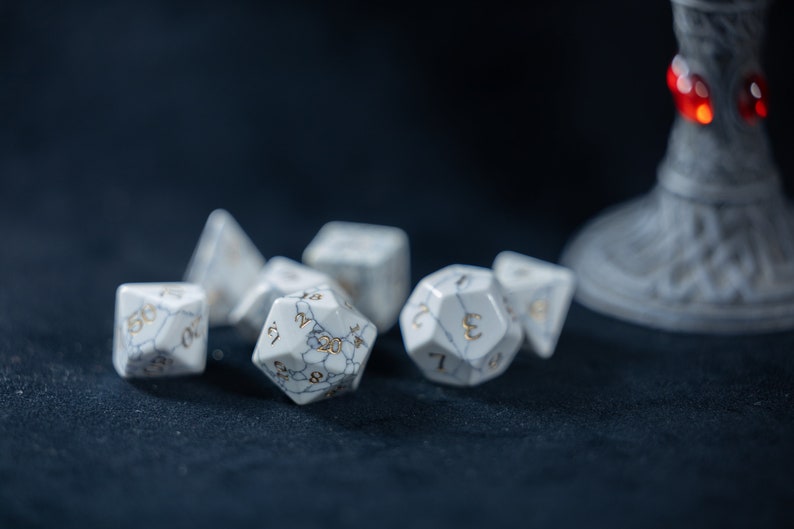 May include: A set of white marble dice with gold numbers. The dice are on a dark blue surface.