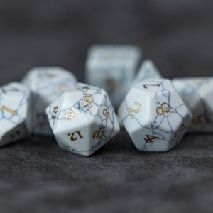 May include: A set of white, marbled dice with gold numbers. The dice are in various shapes and sizes, including a 20-sided die, a 12-sided die, and a 6-sided die.