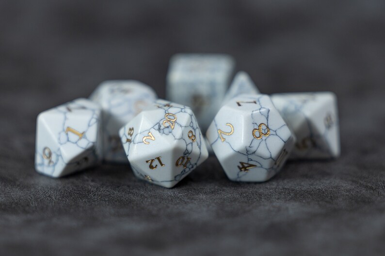May include: A set of white, marbled dice with gold numbers. The dice are in various shapes and sizes, including a 20-sided die, a 12-sided die, and a 6-sided die.