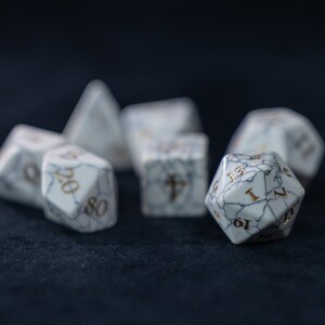 May include: A set of white, marbled dice with gold numbers. The dice are scattered on a dark blue surface.