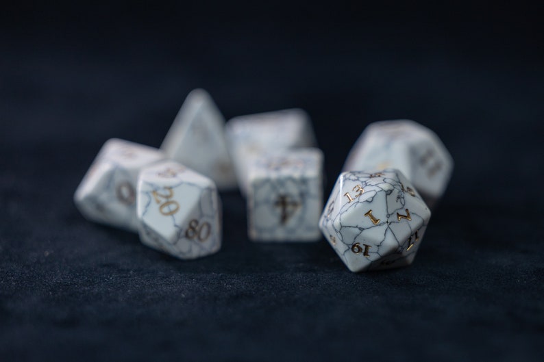 May include: A set of white, marbled dice with gold numbers. The dice are scattered on a dark blue surface.