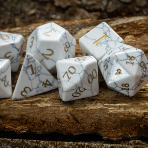 May include: A set of seven white marble dice with gold numbers. The dice are scattered on a wooden surface.