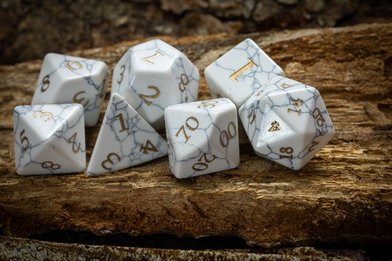 May include: A set of seven white marble dice with gold numbers. The dice are scattered on a wooden surface.