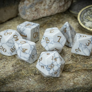 May include: A set of seven white marble dice with gold numbers. The dice are on a light brown, textured surface.