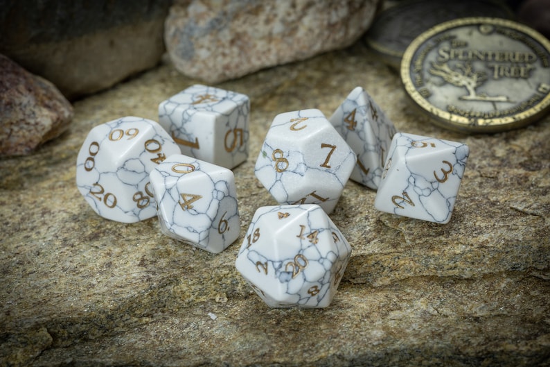 May include: A set of seven white marble dice with gold numbers. The dice are on a light brown, textured surface.
