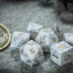 May include: A set of seven white marble dice with gold numbers. The dice are scattered on a gray textured surface.