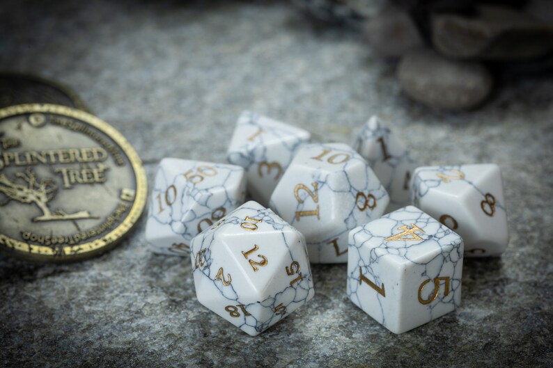 May include: A set of seven white marble dice with gold numbers. The dice are scattered on a gray textured surface.