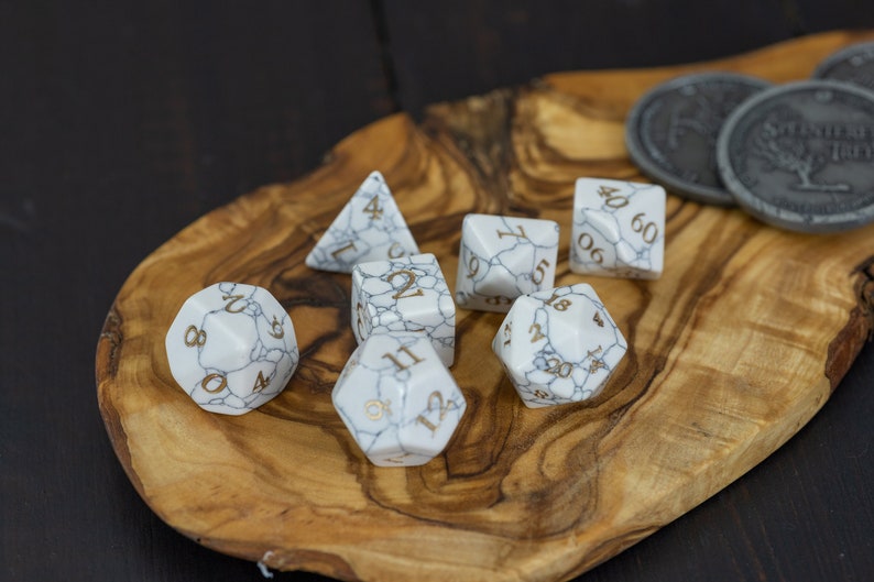 May include: A set of seven white marble dice with gold numbers on a wooden tray. The dice are a d4, d6, d8, d10, d12, and two d20s.
