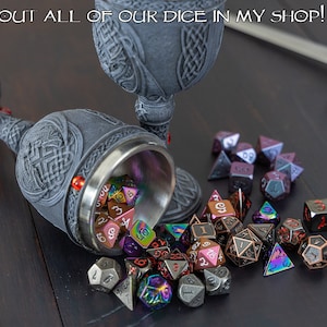 May include: A collection of metal dice in various colors, including iridescent, black, and red, spilling out of a silver chalice with a gray, ornate design. The chalice is resting on a wooden surface.