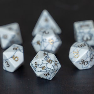 May include: Set of seven white dice with gold numbers. The dice are made of a marbled material with a white and gray pattern. The dice are arranged in a circle with one die in the center.