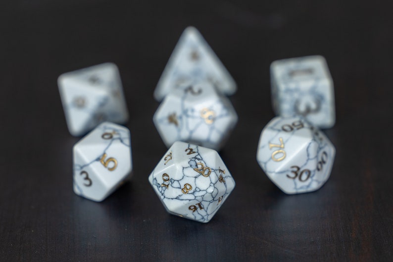 May include: Set of seven white dice with gold numbers. The dice are made of a marbled material with a white and gray pattern. The dice are arranged in a circle with one die in the center.