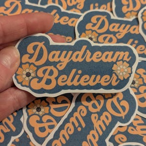Puede incluir: Una colección de pegatinas con fondo azul y texto naranja. Las pegatinas presentan la frase "Daydream Believer" con un diseño floral.