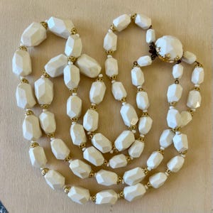 May include: Ivory-colored, multi-strand necklace featuring faceted, geometric beads. The beads are connected by small gold-toned accents. The necklace has a large, round clasp with a gold-toned floral design.