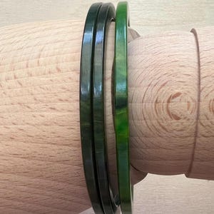 May include: Three smooth, circular bangles with a polished finish. Two of the bangles are a deep, dark forest green, while the third bangle is a lighter, vibrant shade of green with subtle marbled patterns throughout its surface structure.
