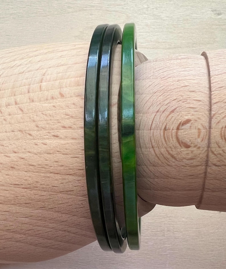 May include: Three smooth, circular bangles with a polished finish. Two of the bangles are a deep, dark forest green, while the third bangle is a lighter, vibrant shade of green with subtle marbled patterns throughout its surface structure.