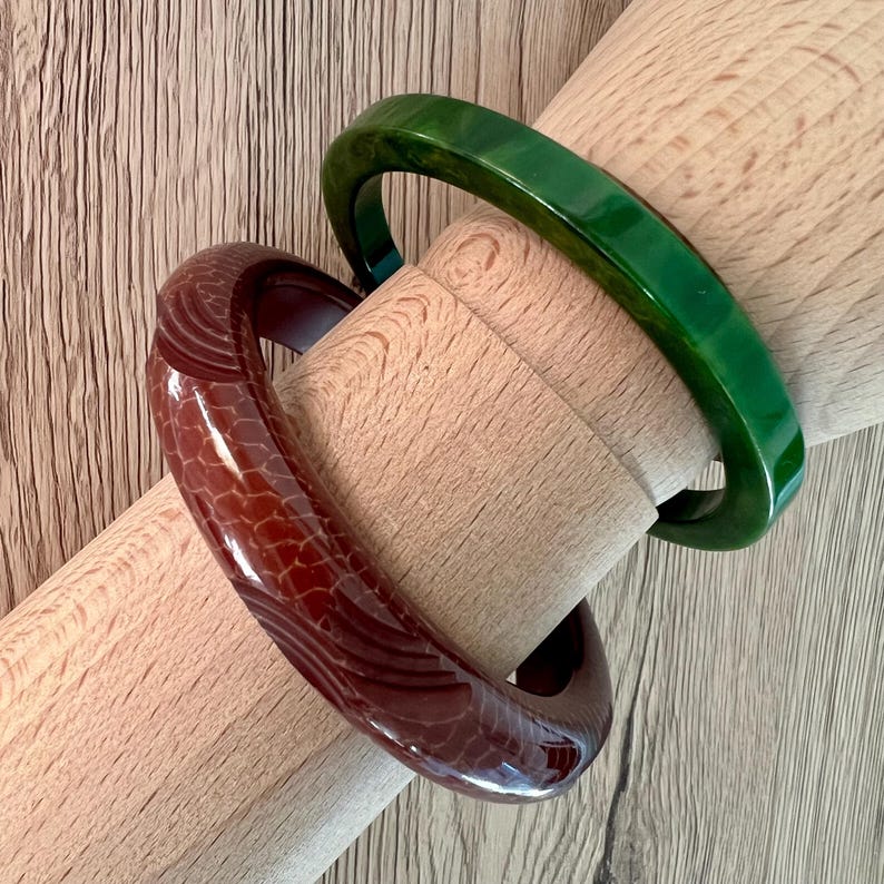 May include: Two circular bangle bracelets. One bracelet is a deep, solid forest green with a smooth, polished finish. The second bracelet is a dark reddish-brown with a textured, scale-like pattern carved into the surface of the material.
