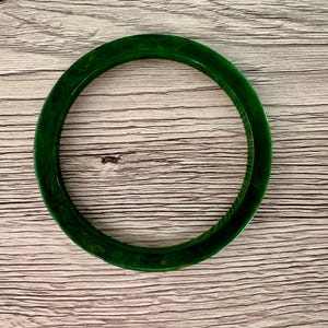 May include: A circular bangle bracelet featuring a deep forest green color with subtle, swirling marbled patterns throughout the smooth, polished surface. The accessory has a rounded profile and a consistent thickness, showcasing a translucent quality.