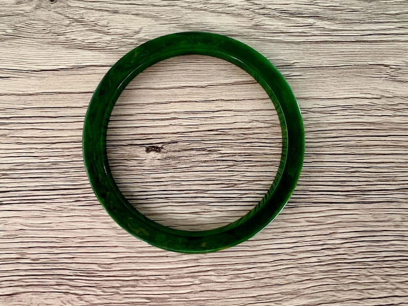 May include: A circular bangle bracelet featuring a deep forest green color with subtle, swirling marbled patterns throughout the smooth, polished surface. The accessory has a rounded profile and a consistent thickness, showcasing a translucent quality.
