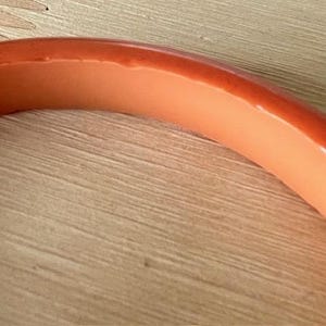 May include: A smooth, curved bangle bracelet featuring a two-tone design with a vibrant orange exterior and a lighter, pale peach-colored interior. The bracelet has a polished, glossy finish and a rounded, solid construction throughout its entire frame.