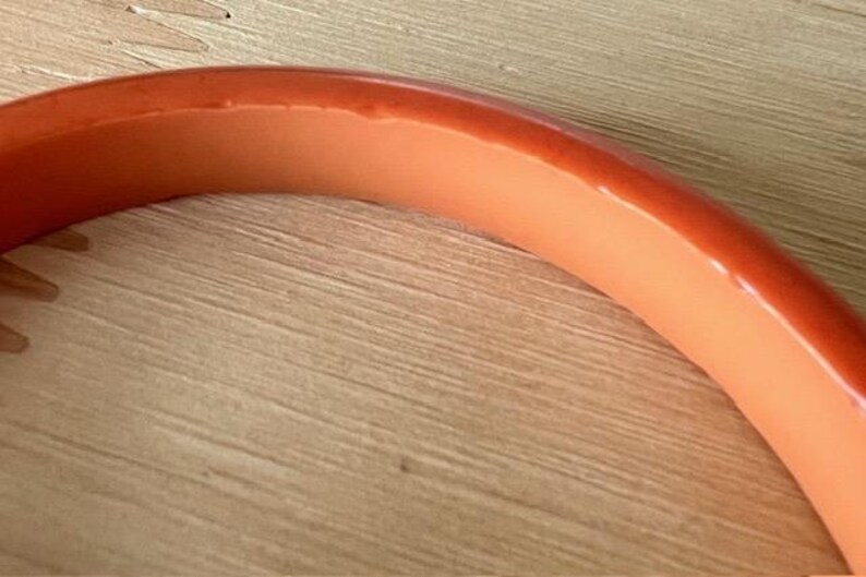 May include: A smooth, curved bangle bracelet featuring a two-tone design with a vibrant orange exterior and a lighter, pale peach-colored interior. The bracelet has a polished, glossy finish and a rounded, solid construction throughout its entire frame.