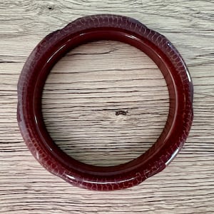 May include: A circular bangle bracelet in a deep, dark red hue. The surface of the bangle features a distinct, raised, and repeating scale-like texture that wraps around the entire circumference of the piece, creating a tactile and patterned appearance.