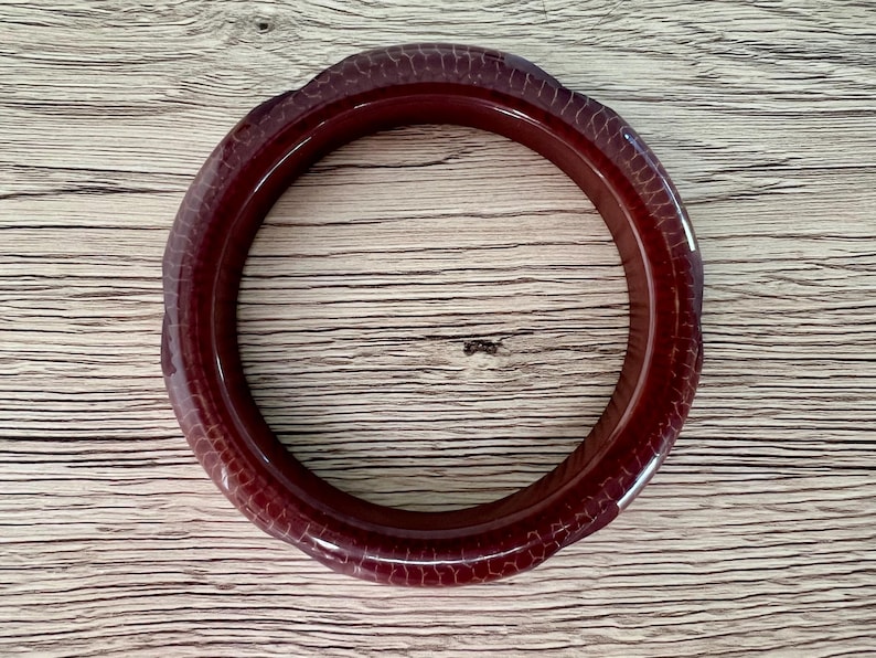 May include: A circular bangle bracelet in a deep, dark red hue. The surface of the bangle features a distinct, raised, and repeating scale-like texture that wraps around the entire circumference of the piece, creating a tactile and patterned appearance.